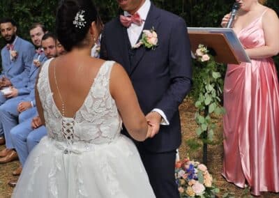 Couple de mariés échangeant un regard pendant leur cérémonie laïque, accompagnés de leur officiante en arrière-plan.